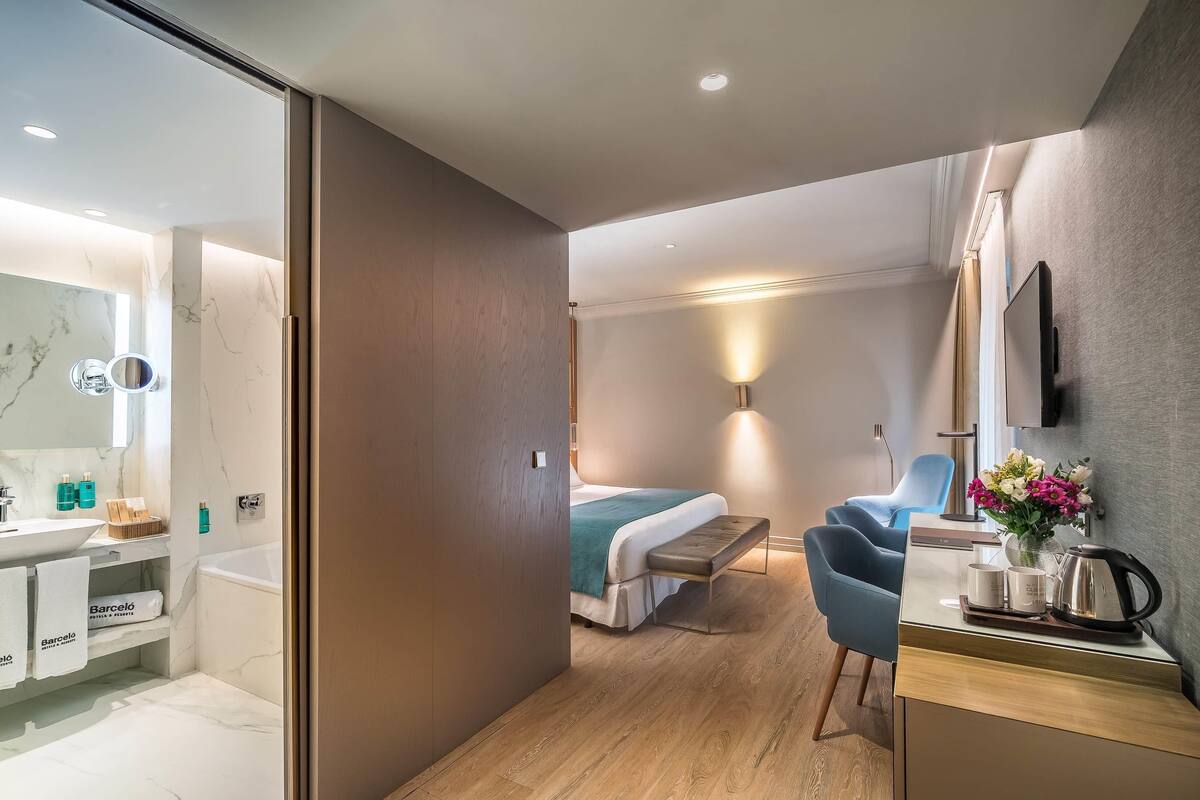 A modern hotel room with a bathroom, a bed, a desk, and a chair.