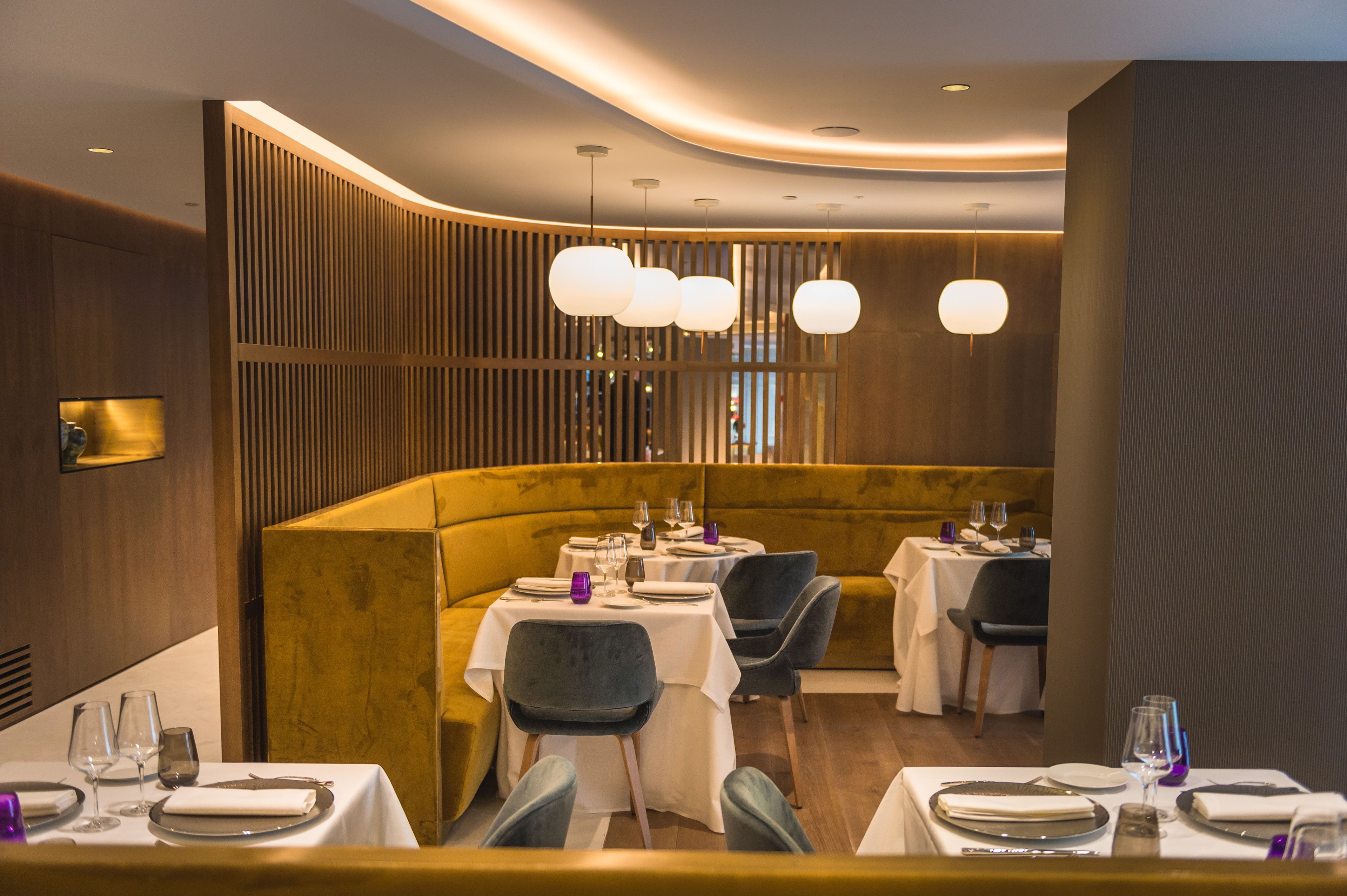 Un restaurante con una decoración moderna, que cuenta con una zona de asientos curva con mesas y sillas, iluminación ambiental y una pared decorativa de madera.