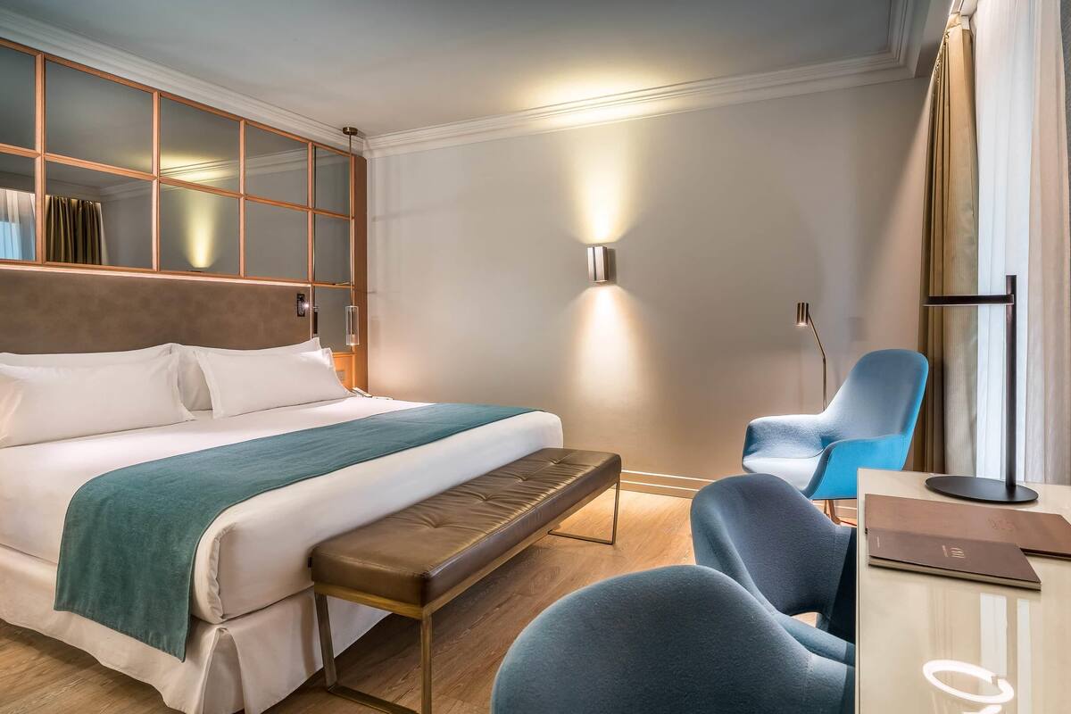 A hotel room with a large bed, a bench, a desk with a laptop, a chair, and a wall-mounted light.