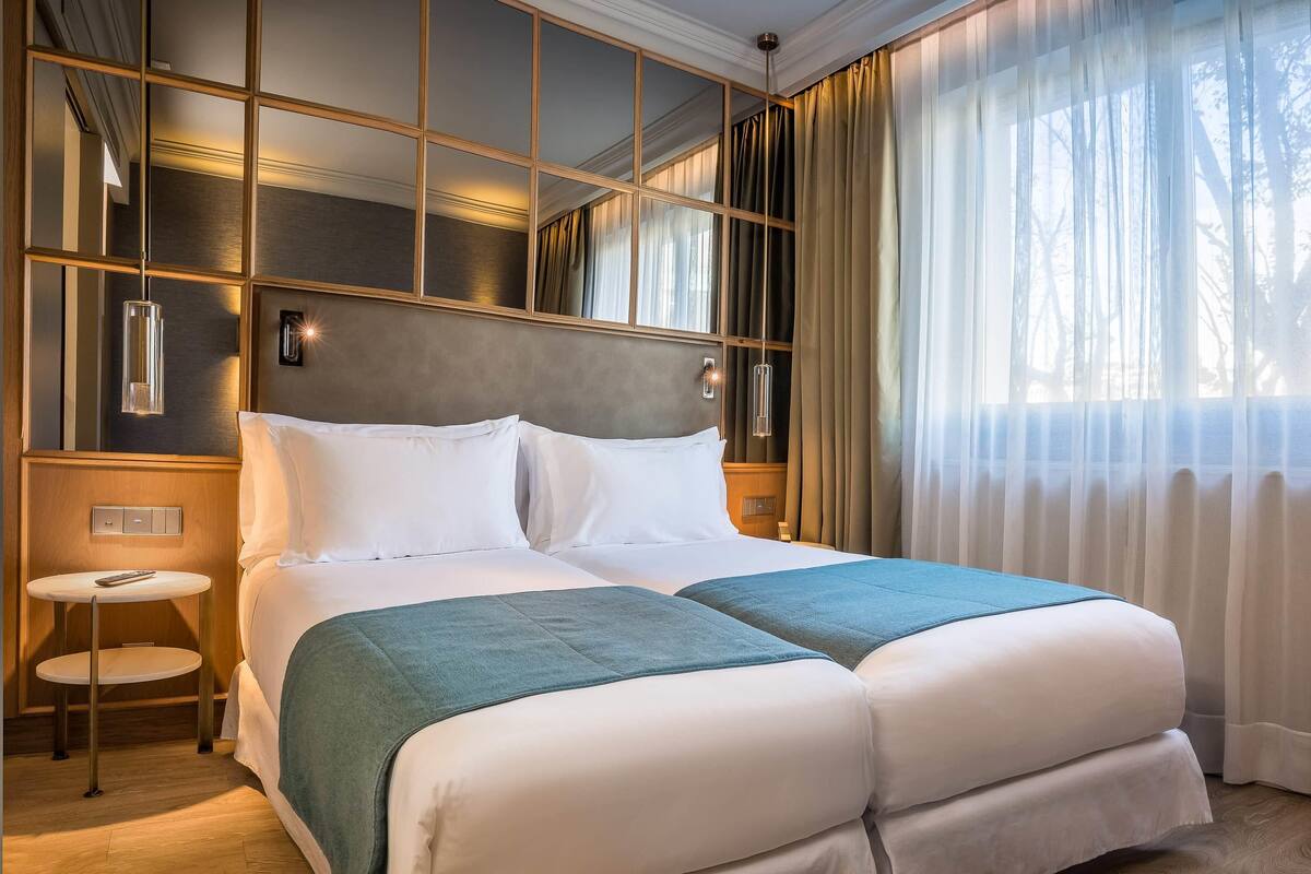 A double bed with white linens and teal throw pillows, a bedside table with a lamp, and a view of trees through sheer curtains.