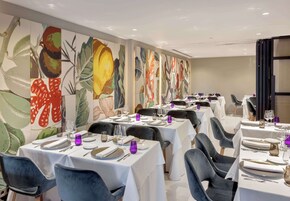 A dining area with tables set for service, featuring a mural with fruit and vegetable designs on the wall.