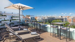 A rooftop terrace with lounge chairs, tables, and an umbrella, overlooking a cityscape.