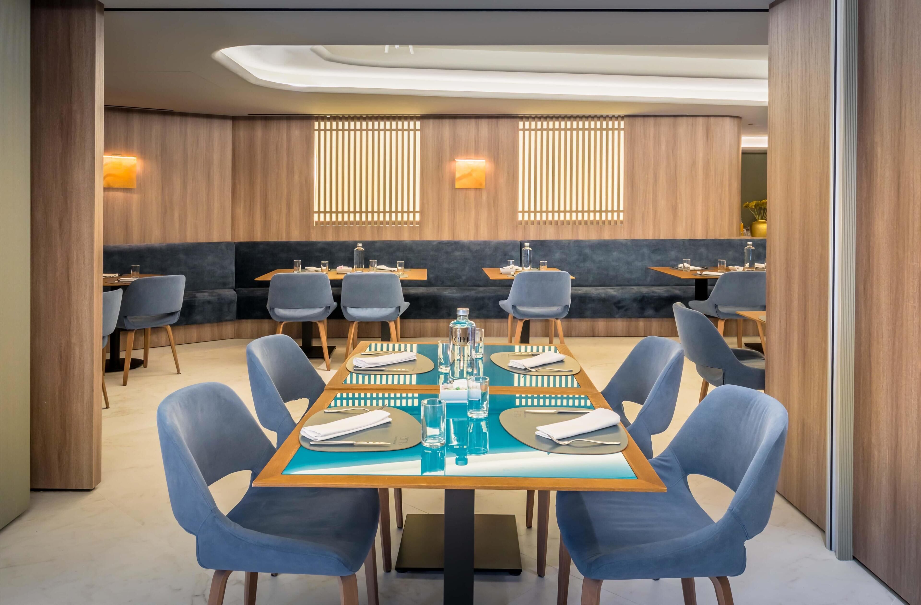 A modern dining area with a long table set for six, surrounded by blue chairs, and a seating area with sofas and chairs in the background.