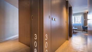A modern hotel hallway with wooden floors and walls, numbered doors, and a dining area visible in the background.