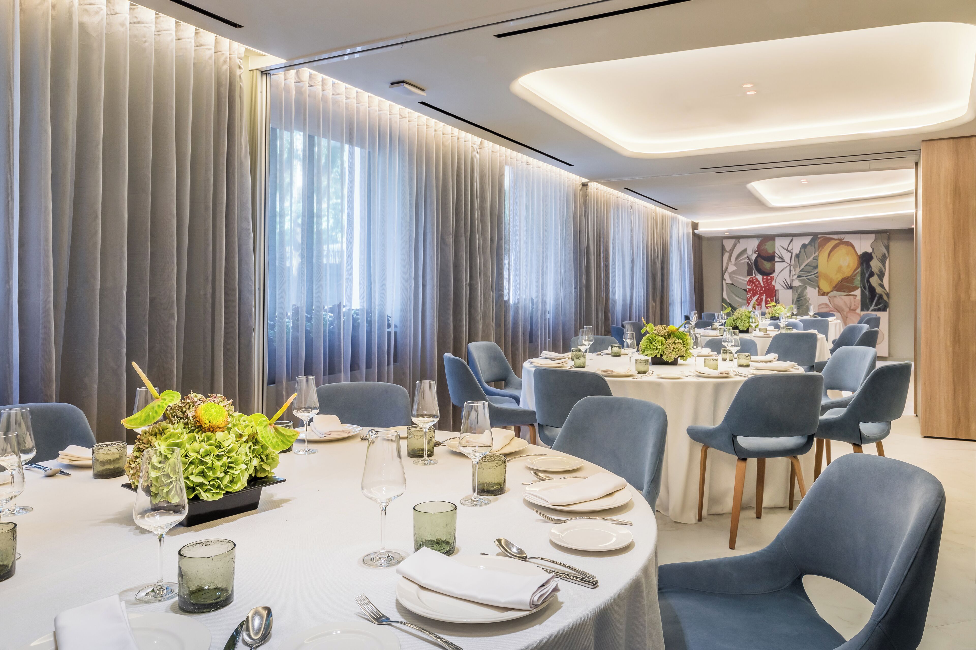 A modern dining area with round tables, blue chairs, and a large window with curtains.