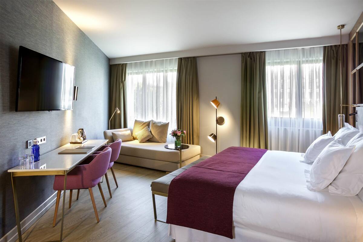 A modern hotel room with a bed, a sofa, a desk, and a TV.
