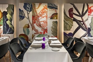 Un'area pranzo con tavoli apparecchiati per quattro persone, ognuno con una tovaglia bianca, sedie nere e bicchieri viola.