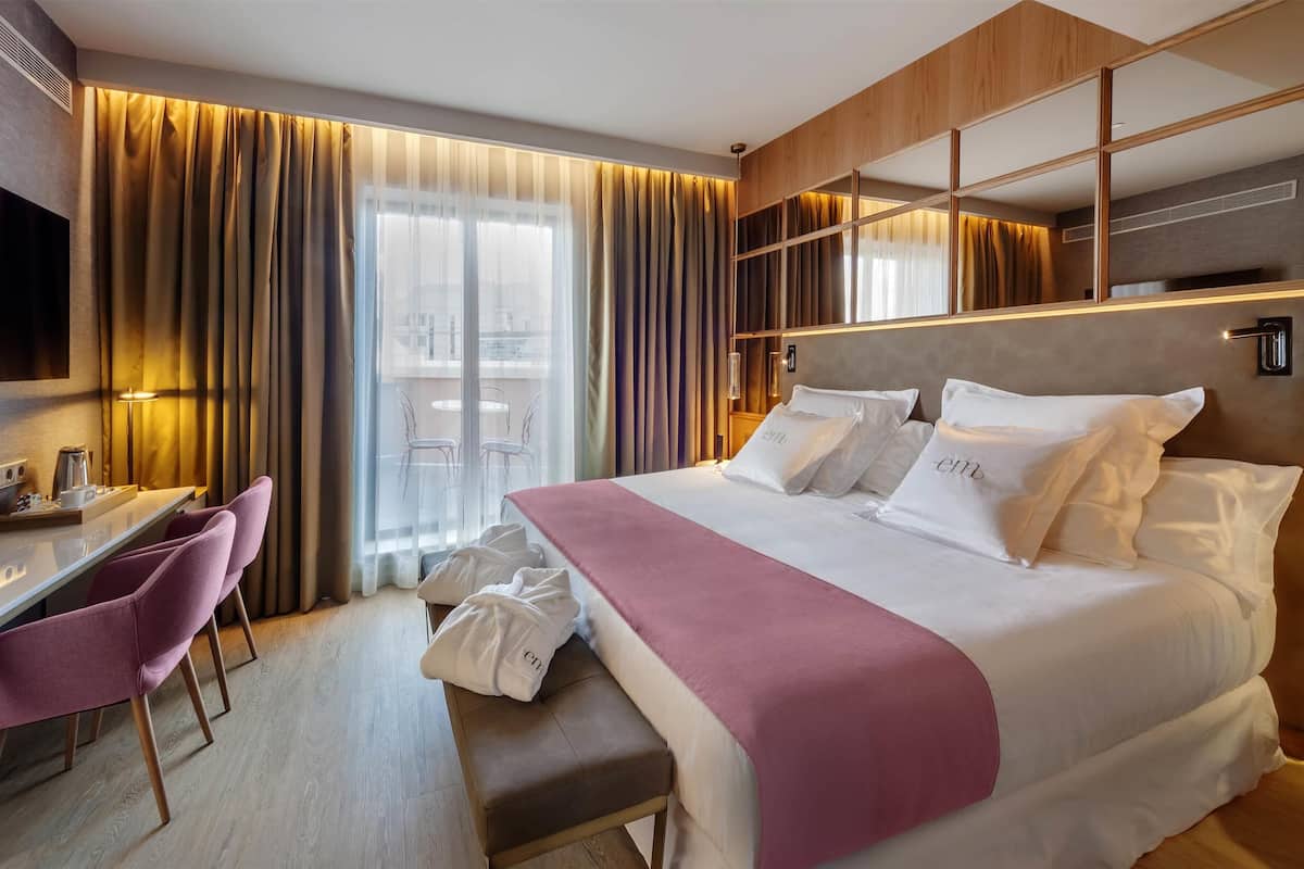 A hotel room with a large bed, a desk with a chair, a bench, and a view of the city through large windows.