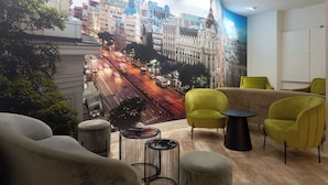 A modern hotel lobby with a mural of a cityscape, comfortable seating, and a clean, well-lit interior. - Hotel Puerta de Toledo (Madrid)