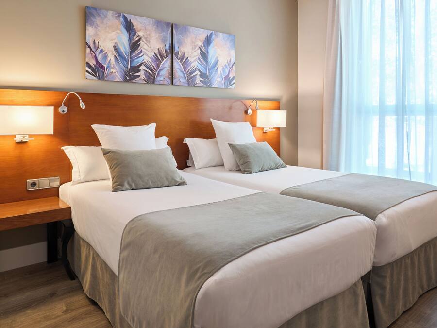 A hotel room with two beds, a wooden headboard, bedside lamps, and a large window with sheer curtains.