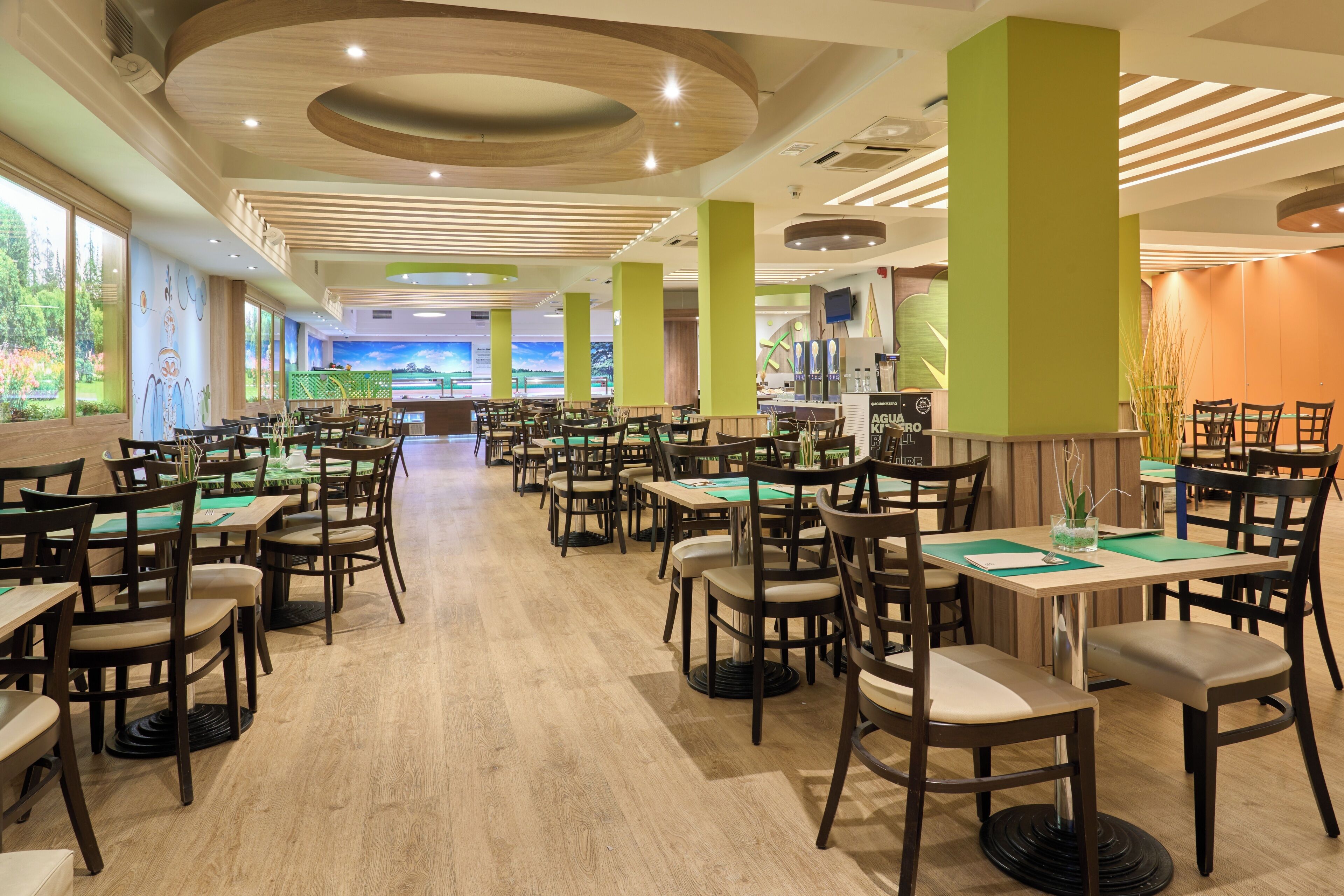 A spacious restaurant with multiple tables and chairs, a wooden floor, and a view of greenery outside.