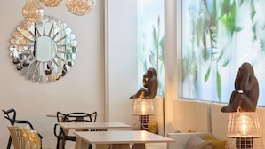 A modern dining area with wooden tables and chairs, decorative light fixtures, and a large window with a nature-themed mural. - Hotel Puerta de Toledo (Madrid)