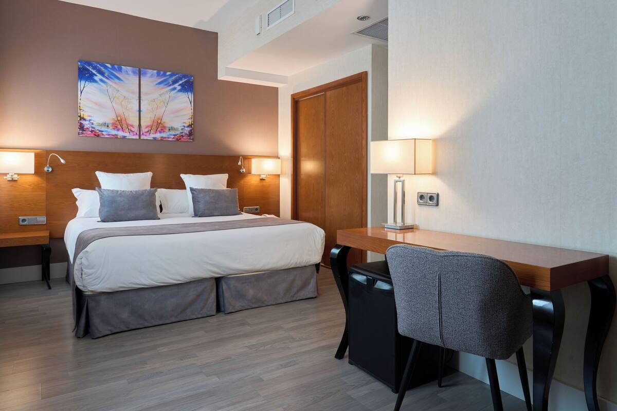 Una habitación de hotel moderna con una cama grande, dos mesitas de noche con lámparas, un escritorio con silla y un armario de madera.