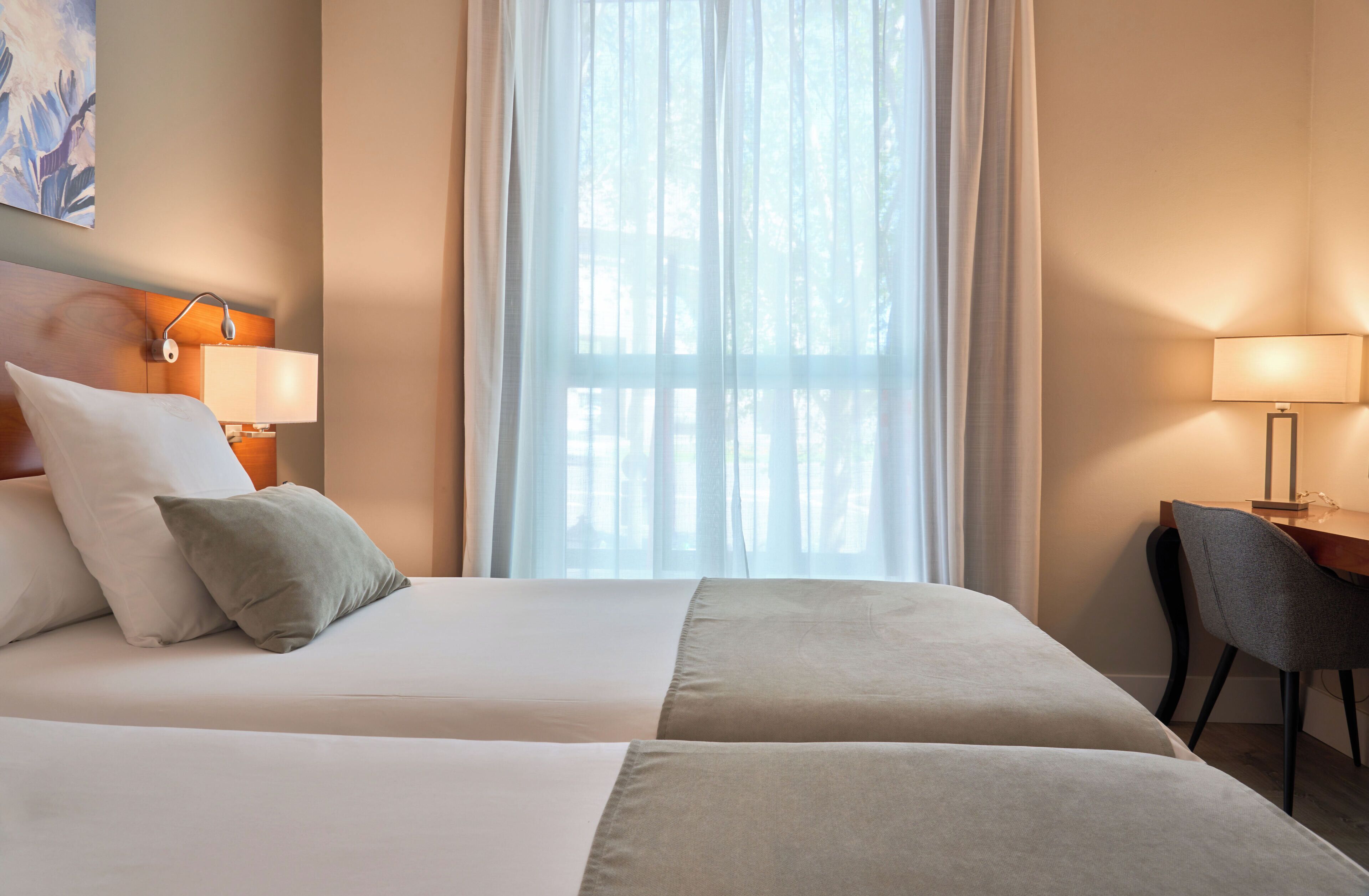 Habitación de hotel con dos camas, un escritorio, una silla y un ventanal con cortinas.