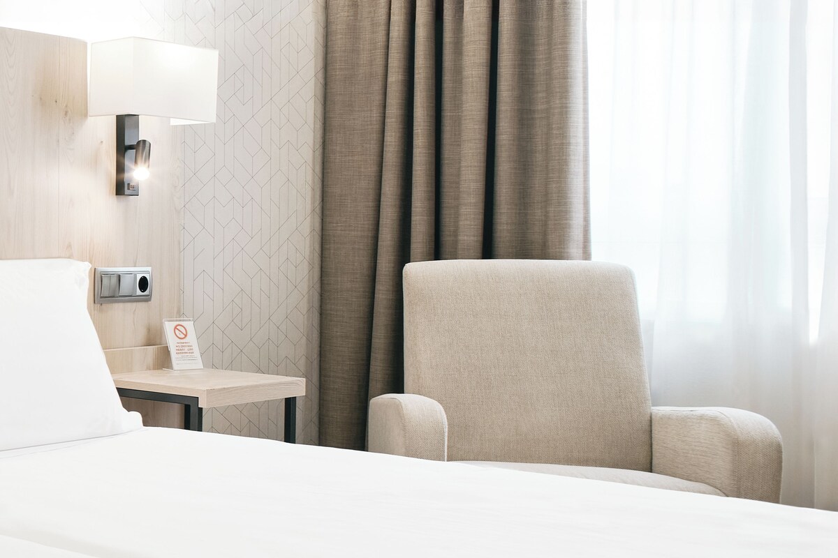 A hotel room with a bed, a chair, a nightstand, a lamp, a sign, and a window with curtains.