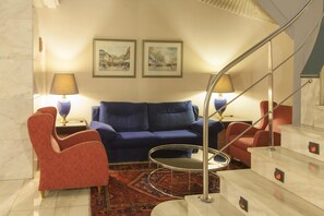 Una sala de estar con un sofá azul, sillones rojos, una mesita de centro y dos lámparas. Hay dos cuadros en la pared.