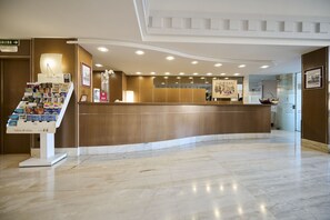 Un área de recepción de hotel con un mostrador curvo, un quiosco de autoservicio con folletos y un cuadro en la pared.