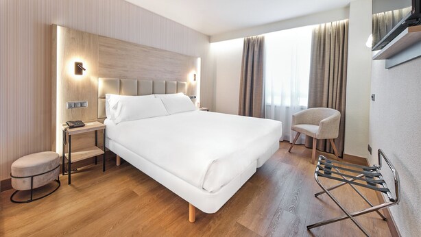 A modern hotel room with a large bed, bedside tables, a chair, a TV, and a window with curtains.
