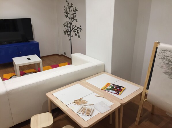 A room with a white sofa, a small white table with a remote control, a wooden table with art supplies, a TV, and a blue cabinet.