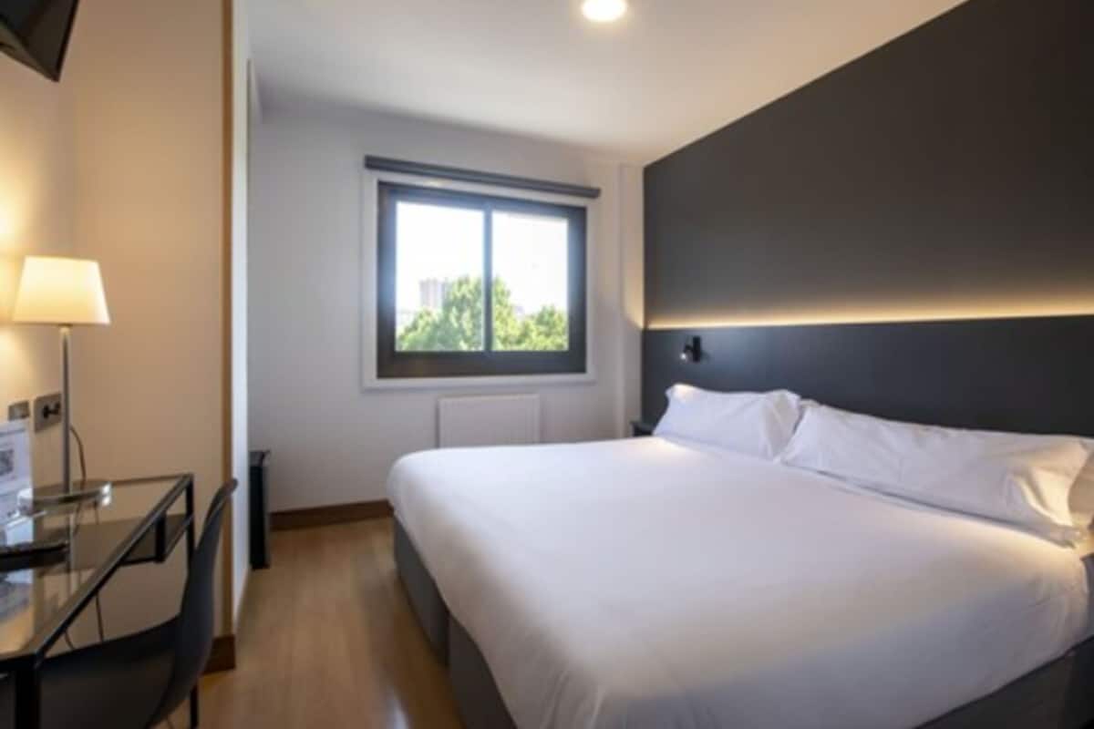Moderni hotellihuone, jossa on suuri sänky, työpöytä ja ikkuna, josta avautuu näkymä vehreään maisemaan.