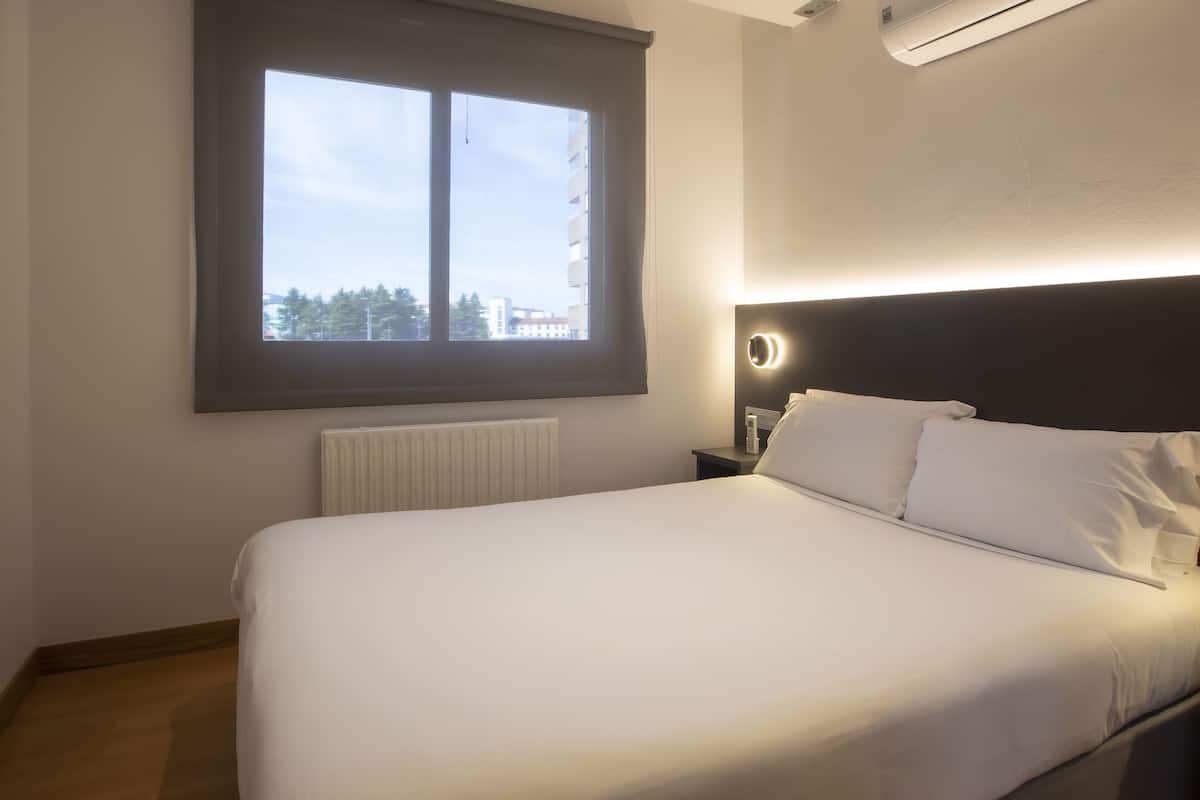 Hotellihuone, jossa on suuri sänky, yöpöytä lampun kanssa, ikkuna, jossa on sälekaihtimet, ja patteri.