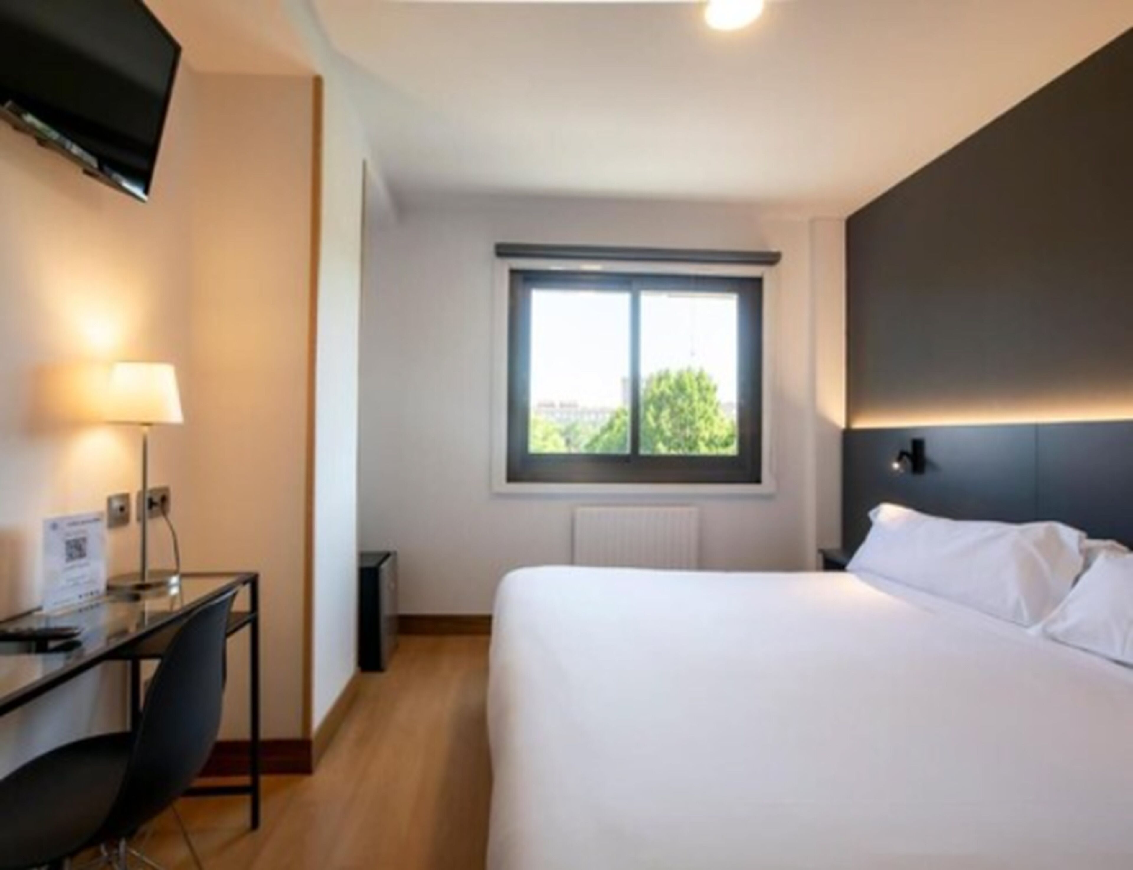 Habitación de hotel con cama, escritorio, silla, televisor montado en la pared y ventana con vistas al verde.