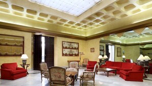 A spacious lobby with a high ceiling, a central seating area with red sofas and floral chairs, and a marble floor.