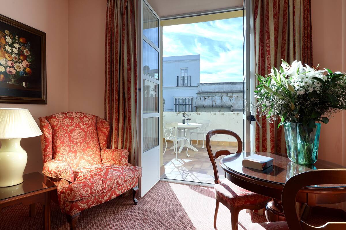 A room with a patterned armchair, a table with a vase of flowers, and a view of a balcony with outdoor furniture.