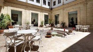 A courtyard with white tables and chairs, potted plants, and a fountain.