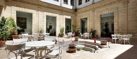 Un patio con mesas y sillas blancas, plantas en macetas y una fuente.