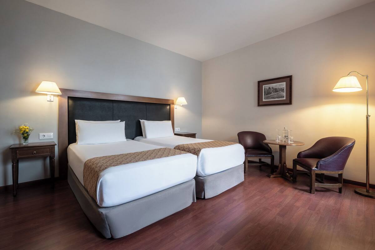 A hotel room with two beds, a desk, a chair, and a lamp.
