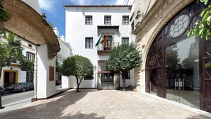 A courtyard with a white building featuring a large arched window and a door, surrounded by trees and a cobblestone path.