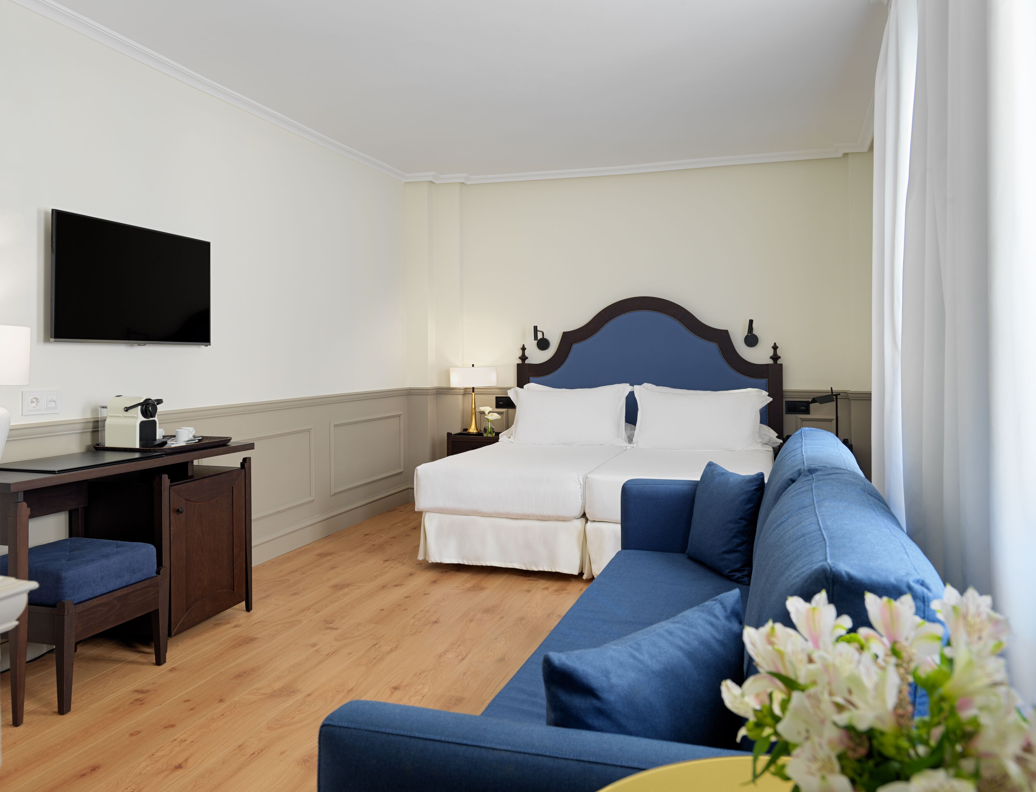 A hotel room with a bed, a desk, a blue sofa, and a TV on the wall.