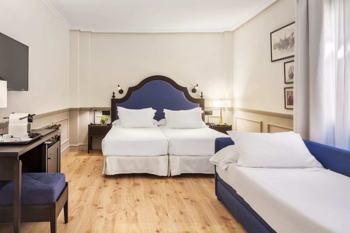 A hotel room with a double bed, a single bed, a desk with a chair, a nightstand with a lamp, and a wall-mounted TV.