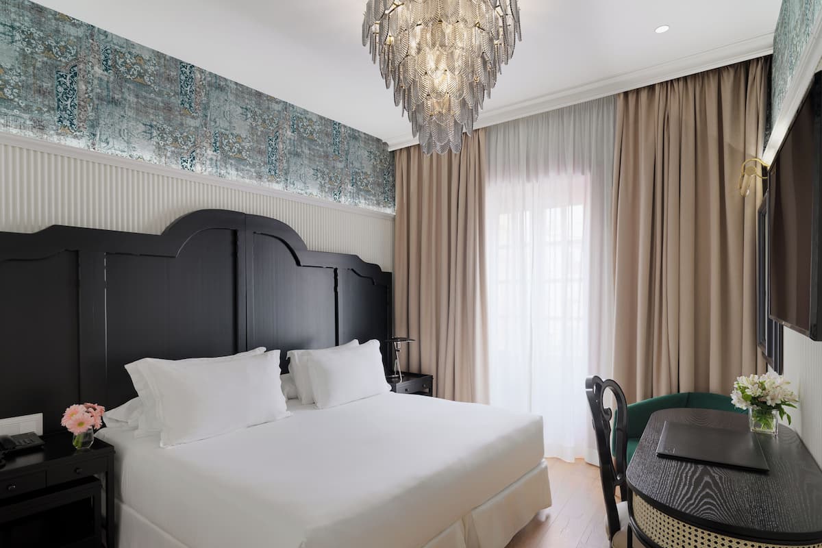 A hotel room with a large bed, a desk, a chair, a chandelier, and a view of the outdoors.