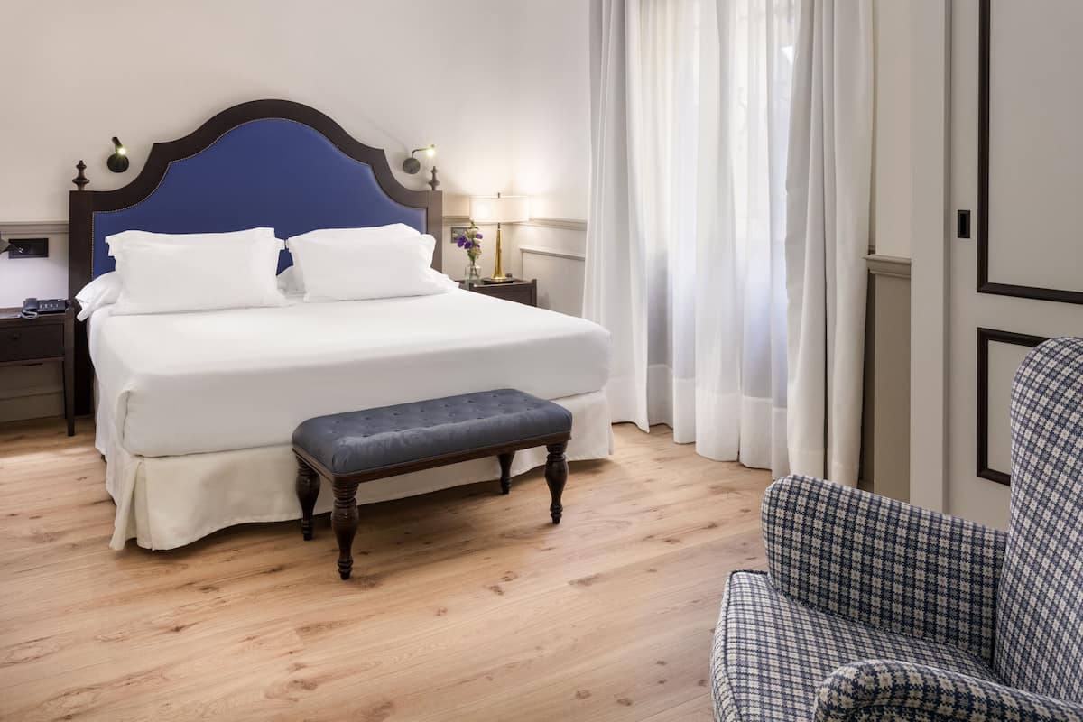 A neatly made bed with white linens and a dark headboard, a wooden bench at the foot of the bed, and a checkered armchair.