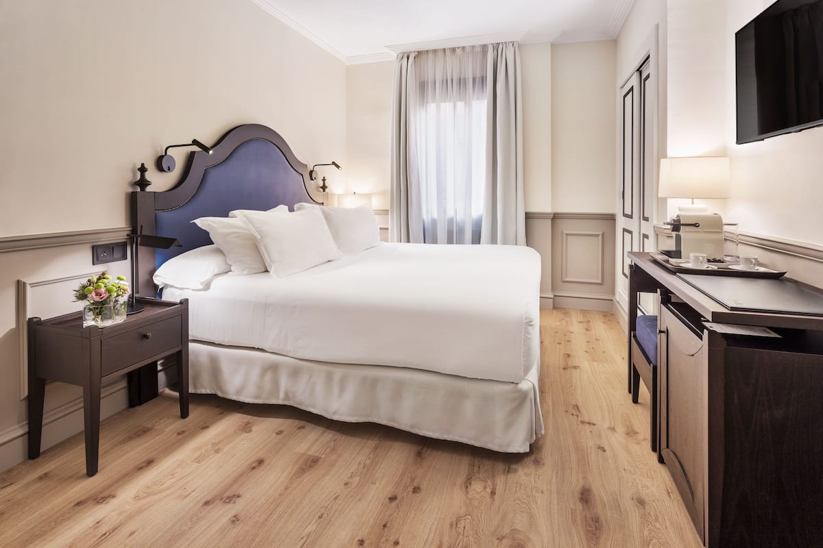 A hotel room with a large bed, a bedside table with a lamp, a desk with a chair, and a TV mounted on the wall.