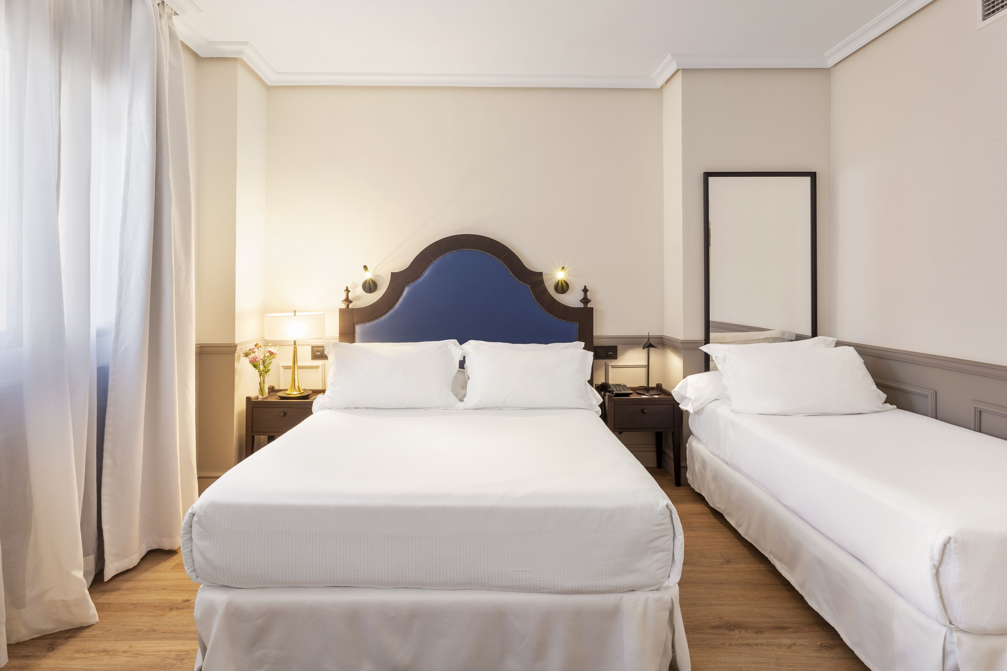 Habitación de hotel con dos camas individuales, una mesita de noche con lámpara, un espejo y una ventana con cortinas.