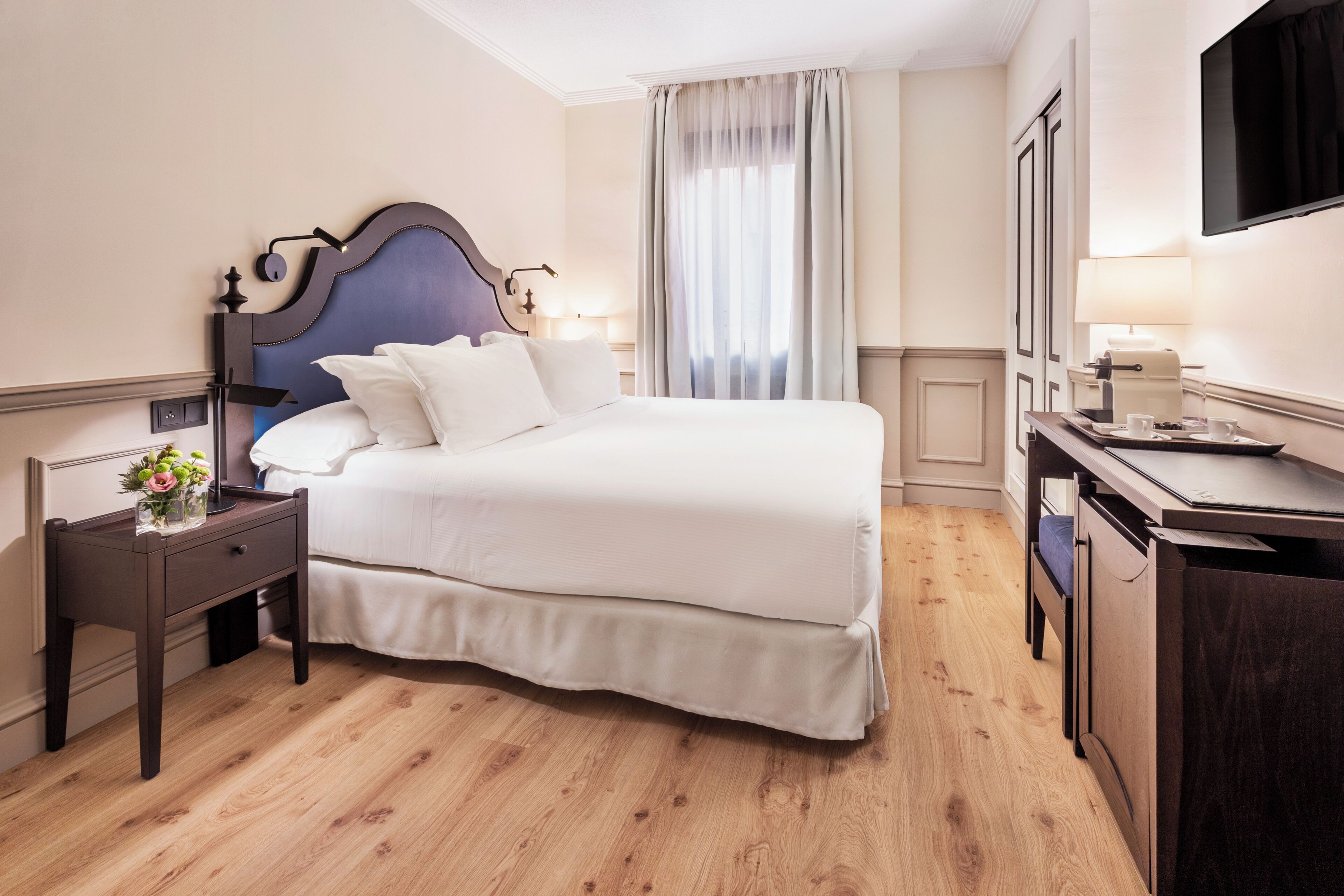 A hotel room with a large bed, a bedside table with a lamp, a desk with a chair, and a TV mounted on the wall.