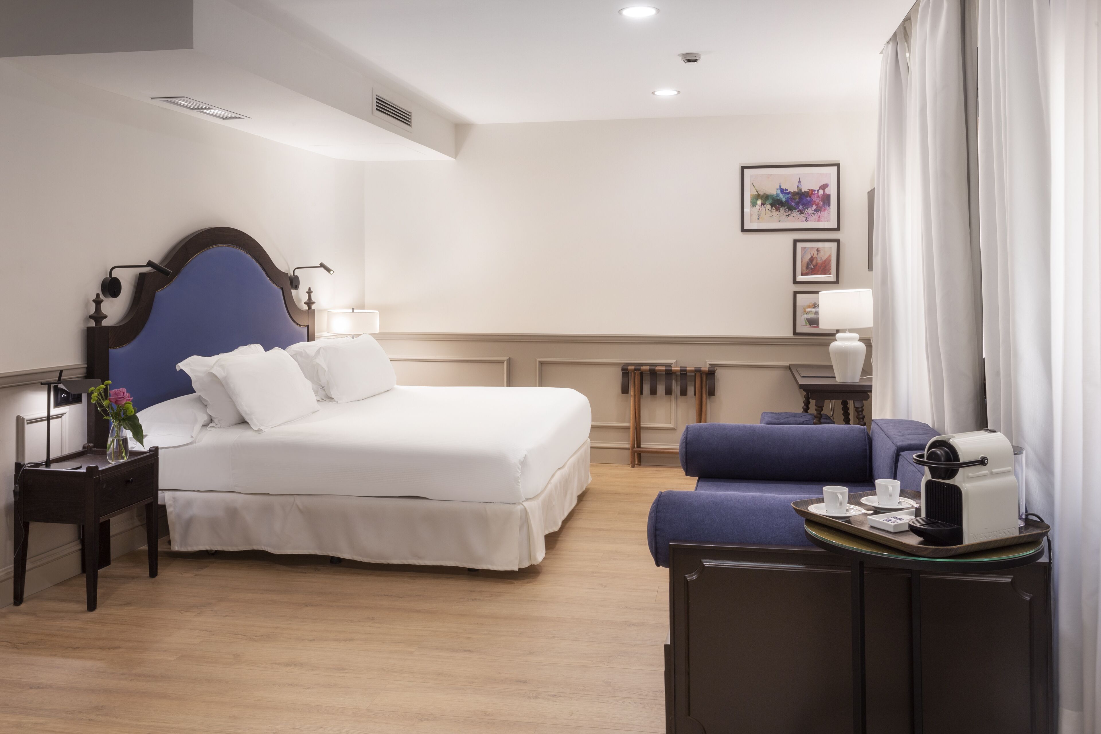 Una habitación de hotel con una cama grande, un sofá pequeño, una mesita de noche con lámpara, una bandeja con tazas y cafetera, y un cuadro en la pared.