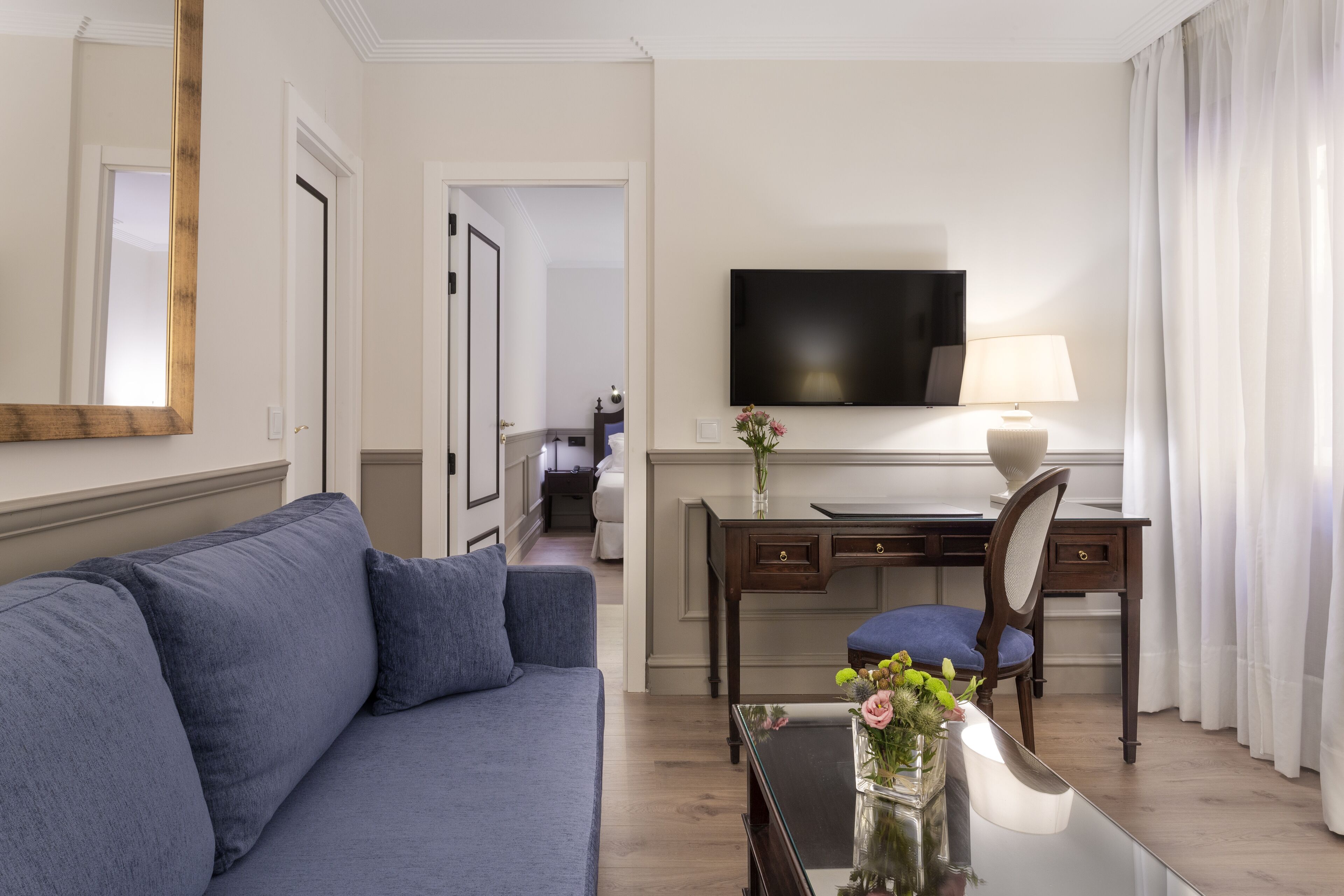 A hotel room with a blue sofa, a desk with a chair, a flat-screen TV, and a vase with flowers.
