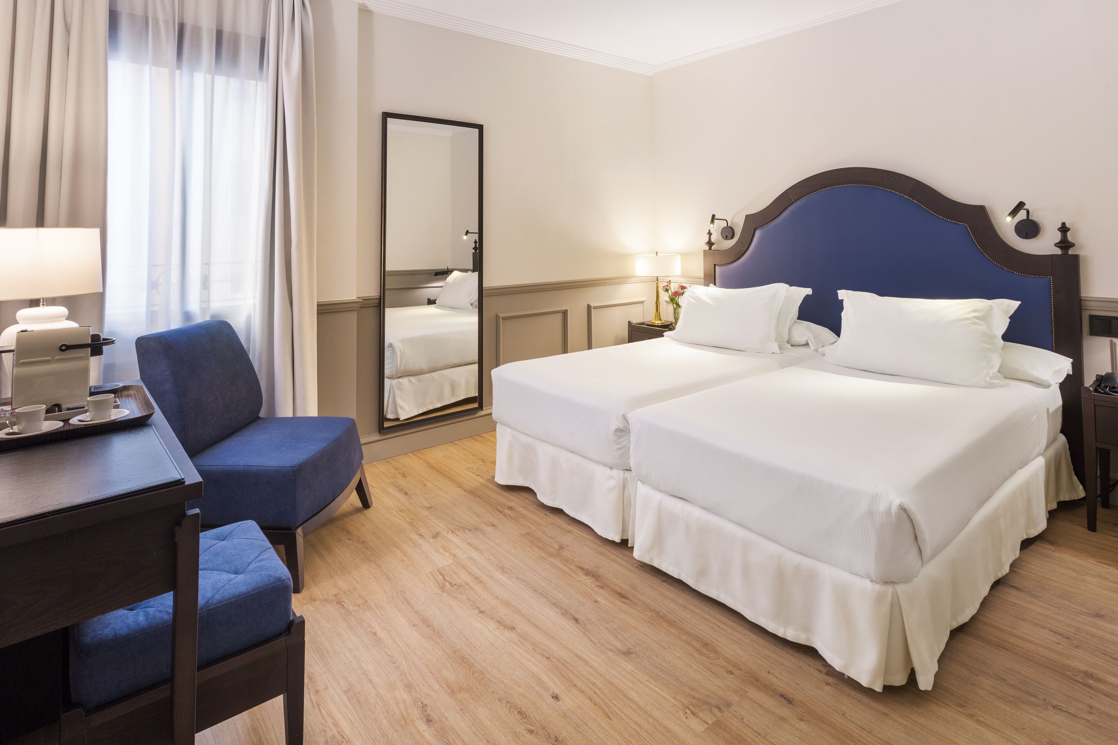 A hotel room with two beds, a desk with a chair, a mirror, and a window with curtains.