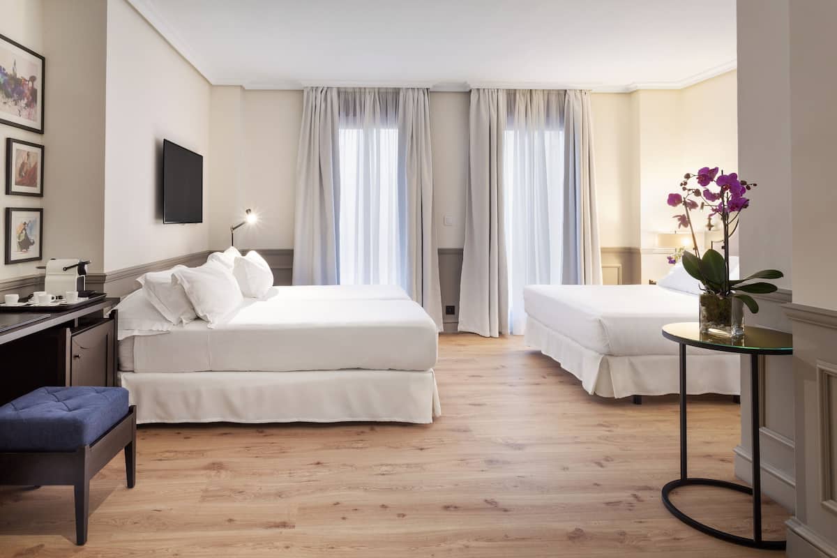 A hotel room with two beds, a desk with a chair, a small table with a vase of flowers, and a flat-screen TV mounted on the wall.