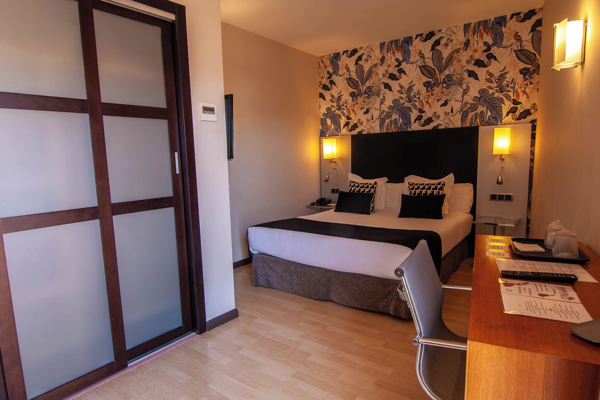 A hotel room with a bed, a desk with a chair, a desk lamp, and a wall-mounted light.