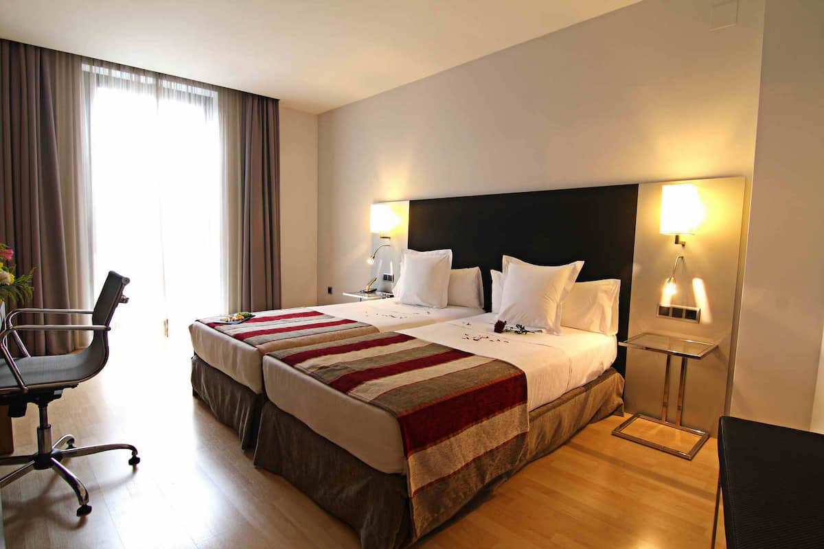 Une chambre d’hôtel avec un grand lit, deux tables de chevet avec des lampes, un bureau avec une chaise et une fenêtre avec des rideaux.