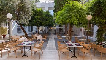 An outdoor cafe with tables and chairs arranged in rows, surrounded by trees and a building in the background.