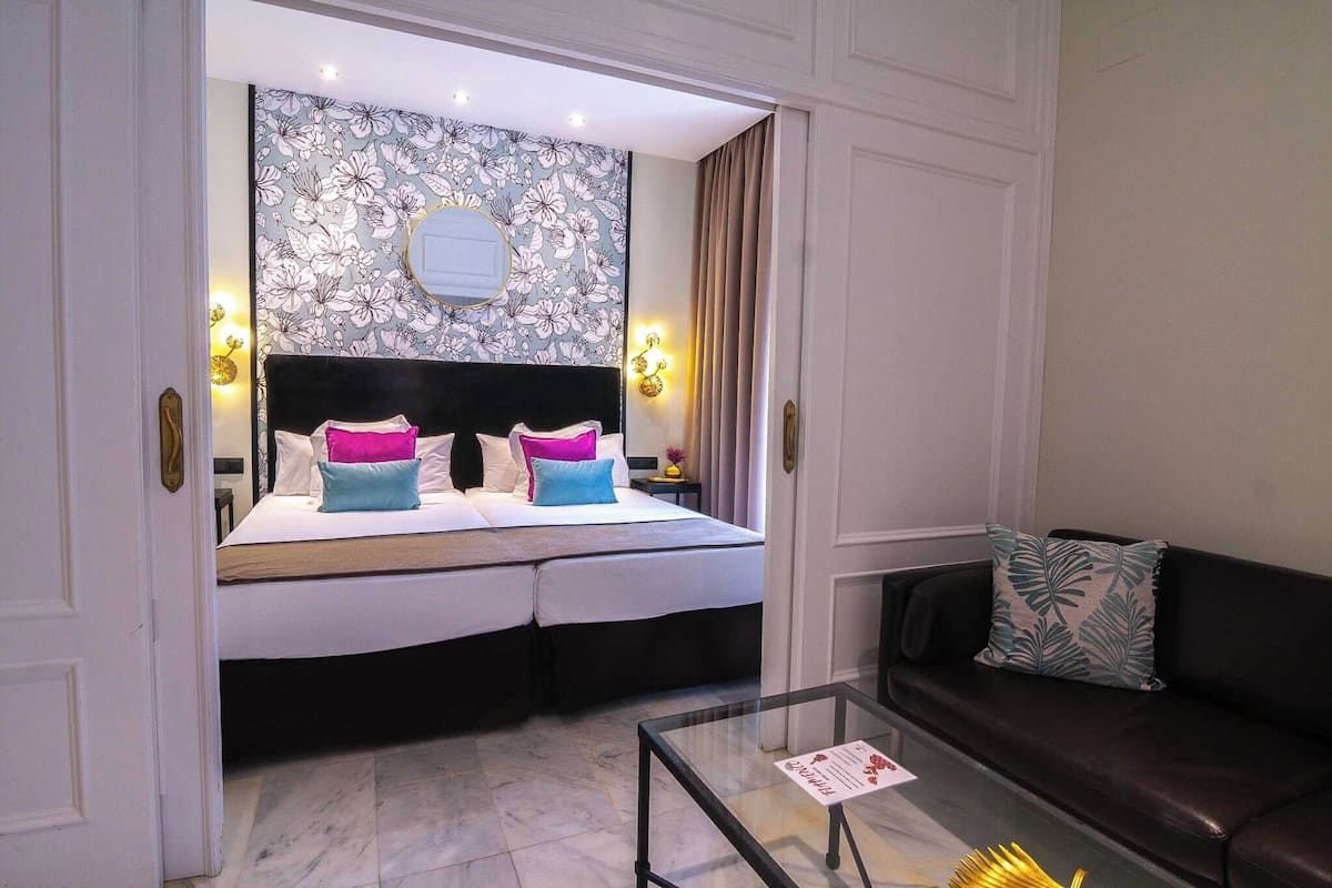 Une chambre d’hôtel avec un lit, un canapé, une table basse en verre et un mur orné d’un motif floral.