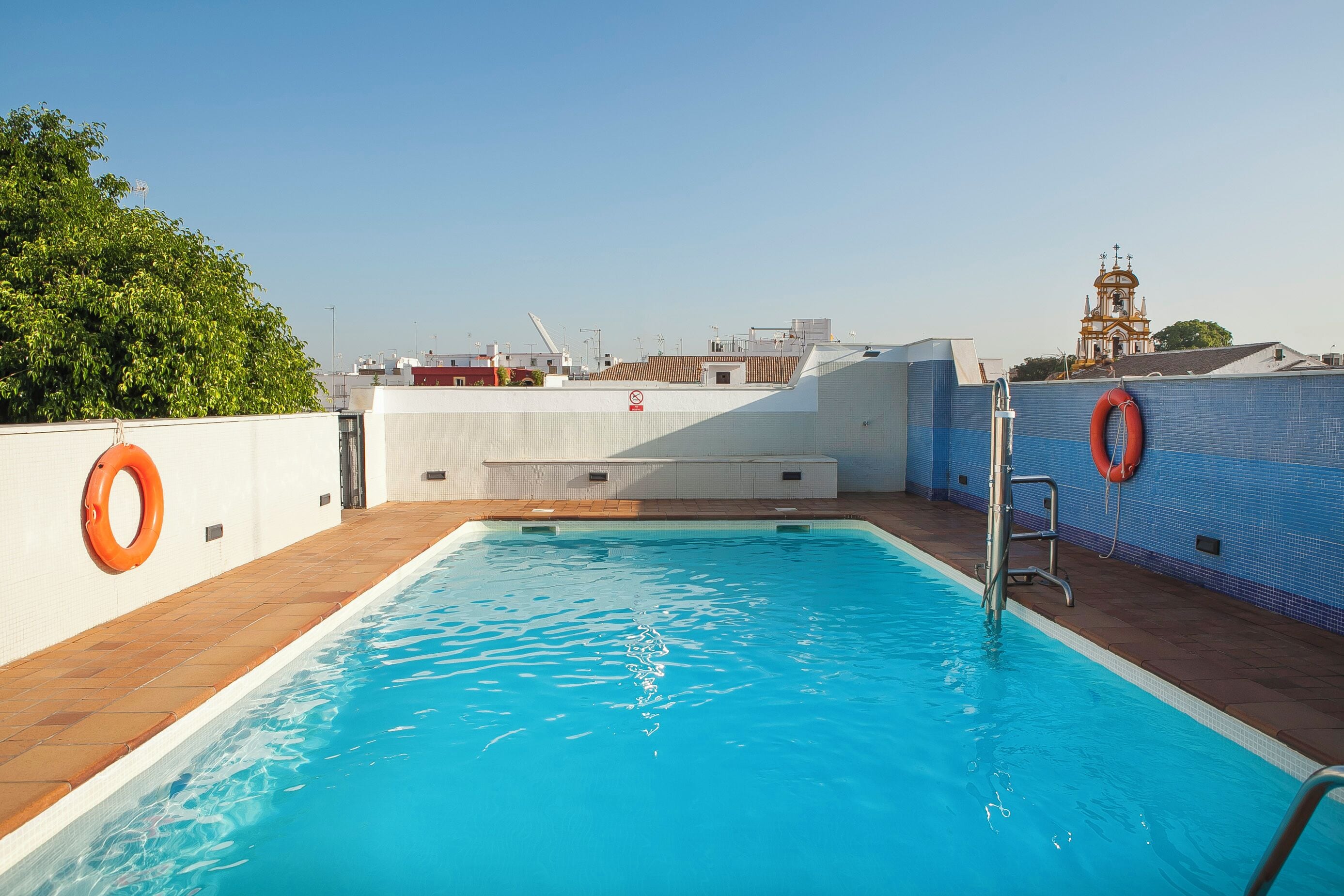 A rooftop swimming pool with clear blue water, a lifebuoy, and a view of a church tower in the background.