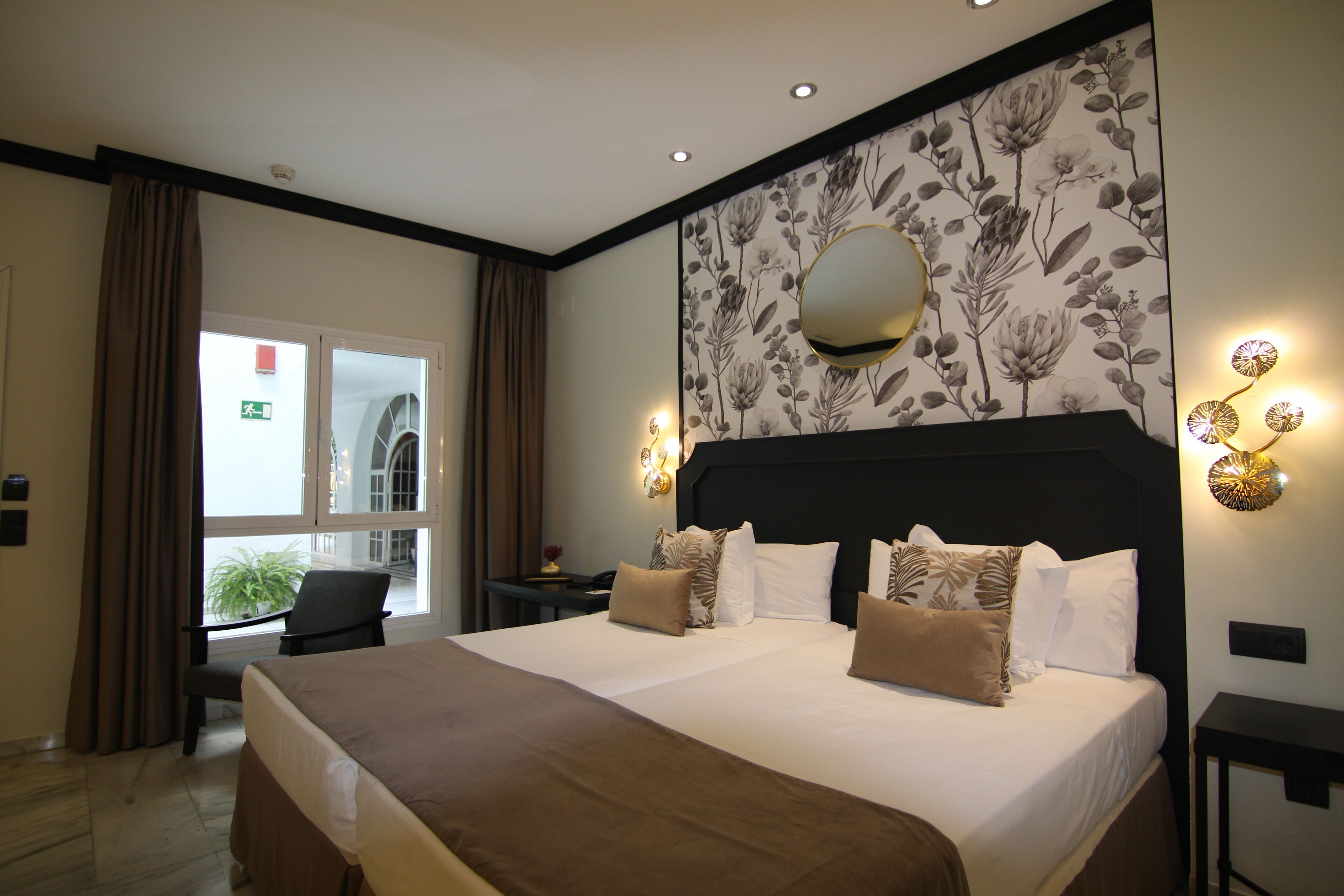 Habitación de hotel con una cama grande, un escritorio, una silla, un ventanal con cortinas, y una pared con papel tapiz floral y un espejo.