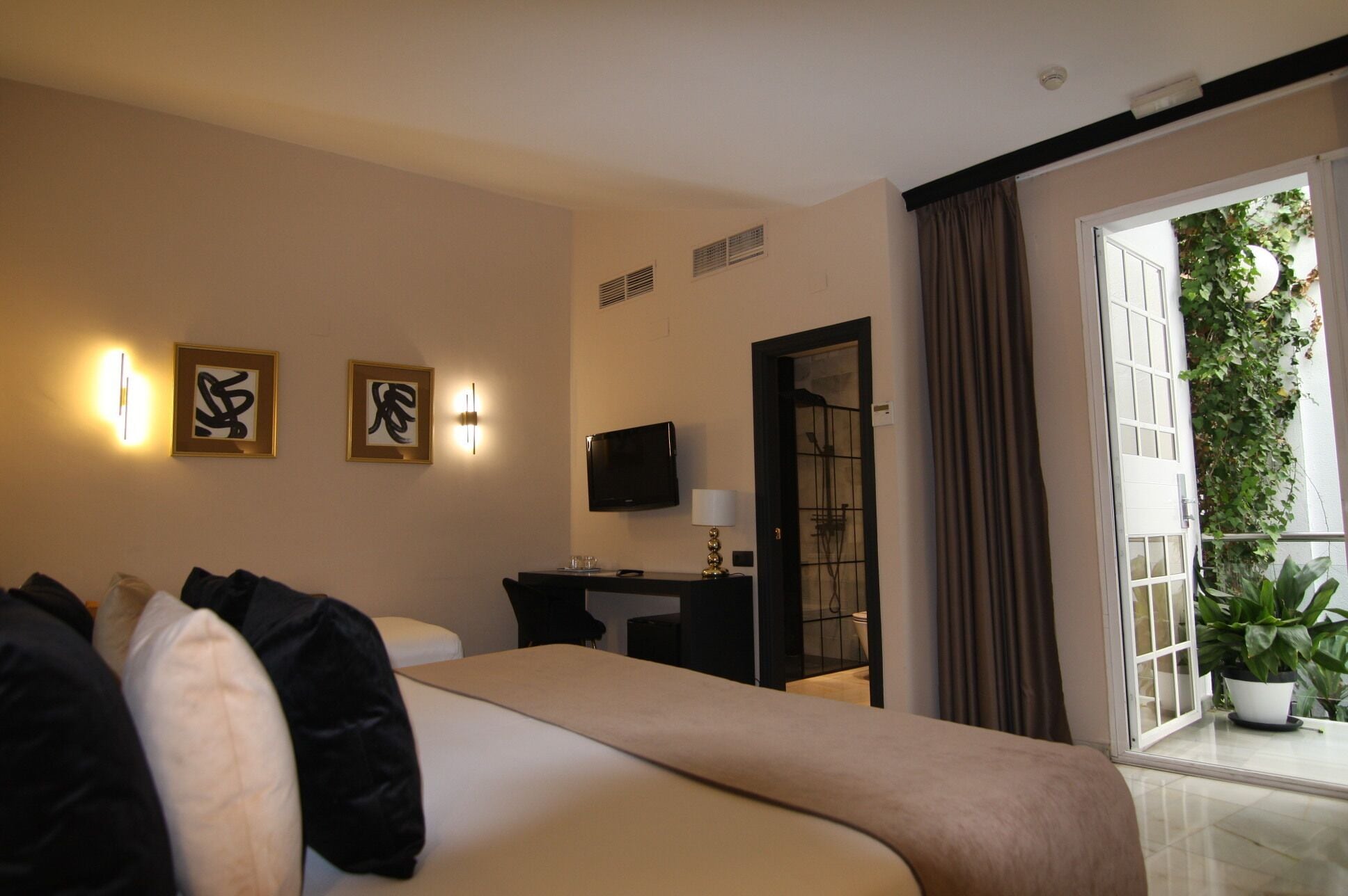 A hotel room with a bed, a desk with a TV, and a balcony with plants.